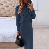 Frau in blauem Strickkleid mit Sternenmuster, hält schwarze Handtasche, modernes Schlafzimmer.